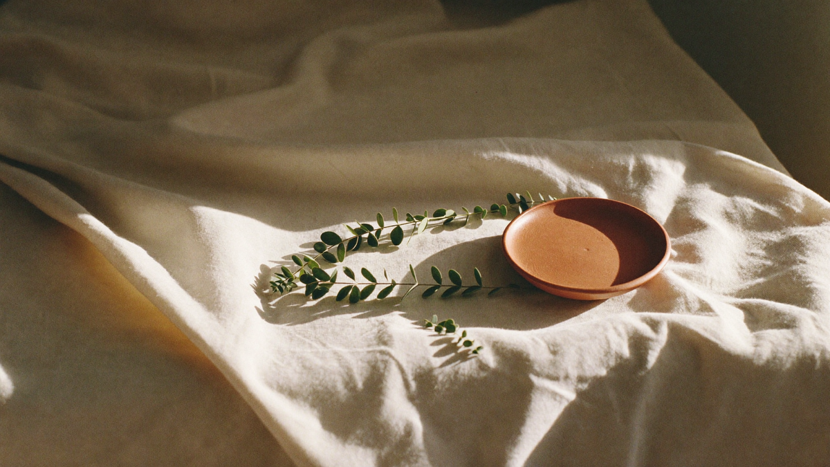 Warm morning light across cream linen with sage eucalyptus sprigs and a terracotta dish
